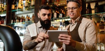 Mid adult barista and his young coworker using touchpad while working in a pub.
